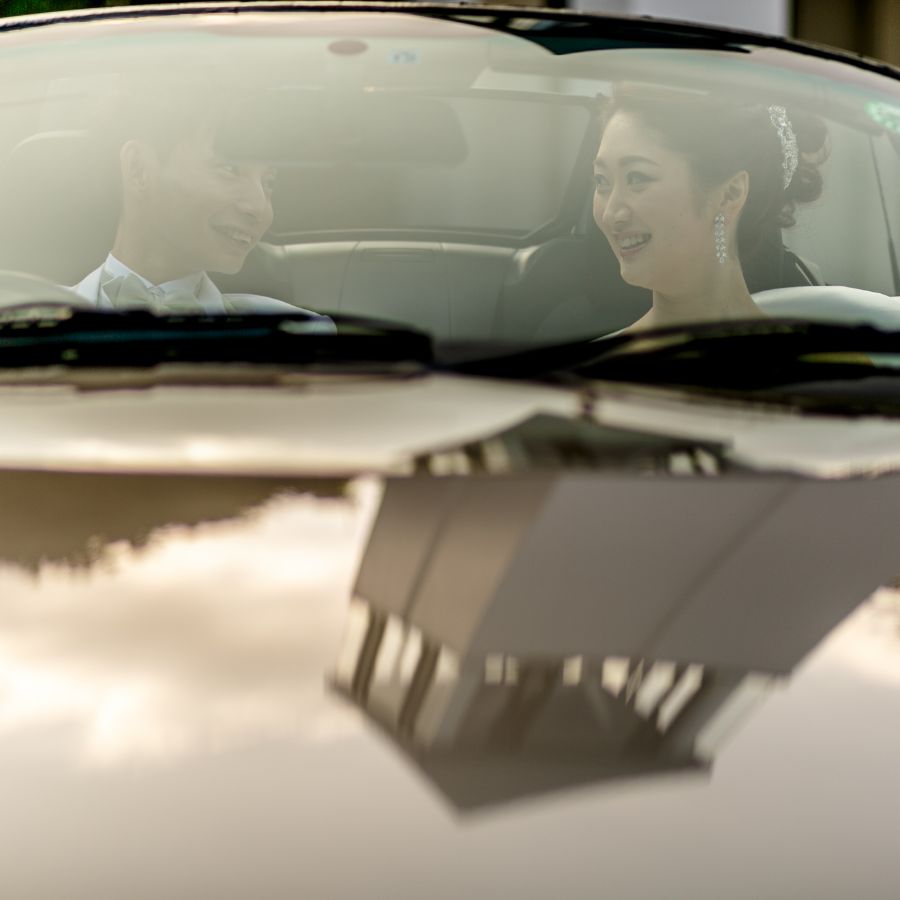 新郎新婦オープンカーに乗り、ボンネットにはチャペルのカリオンが映りこんでいる